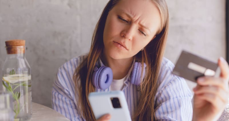 Tüdruk, kes kannab kaelas kõrvaklappe, vaatab käes olevasse telefoni ja hoiab teises käes pangakaarti.