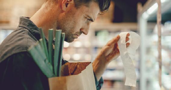 Mees seisab poes külmkapi ees ning hoiab ühe käega paberkotti ning teise käega ostutšekki, mida ta vaatab.