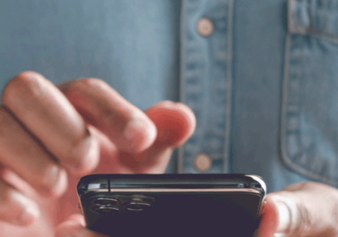 A person in a denim shirt holds a smartphone and taps the screen with their finger. A chat bubble pops up on the screen with the text 